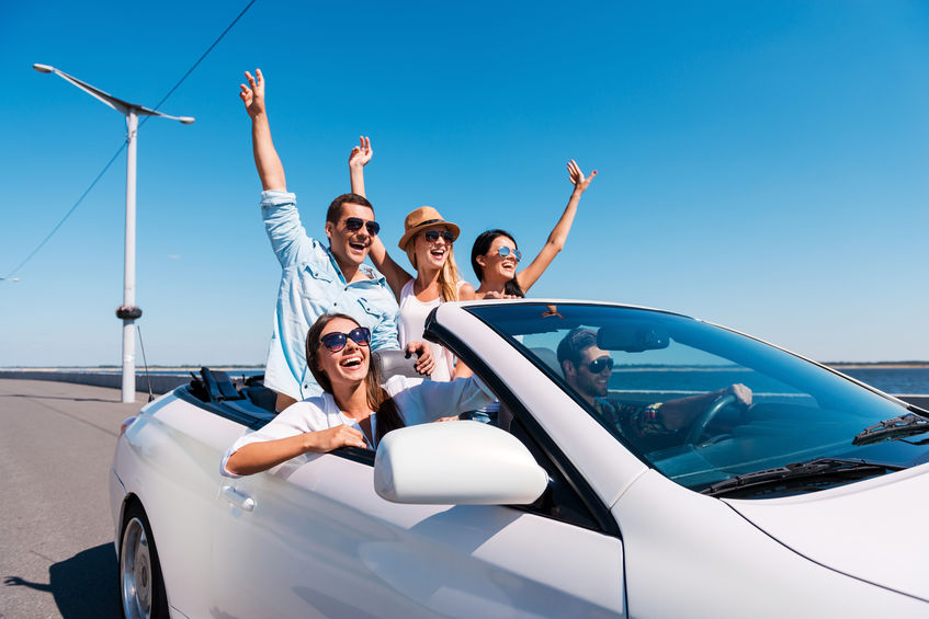 Groupe d'amis enthousiastes dans une décapotable blanche lors d'un road trip dans l'Est du Canada, bras en l'air et sourires radieux sous un ciel bleu éclatant